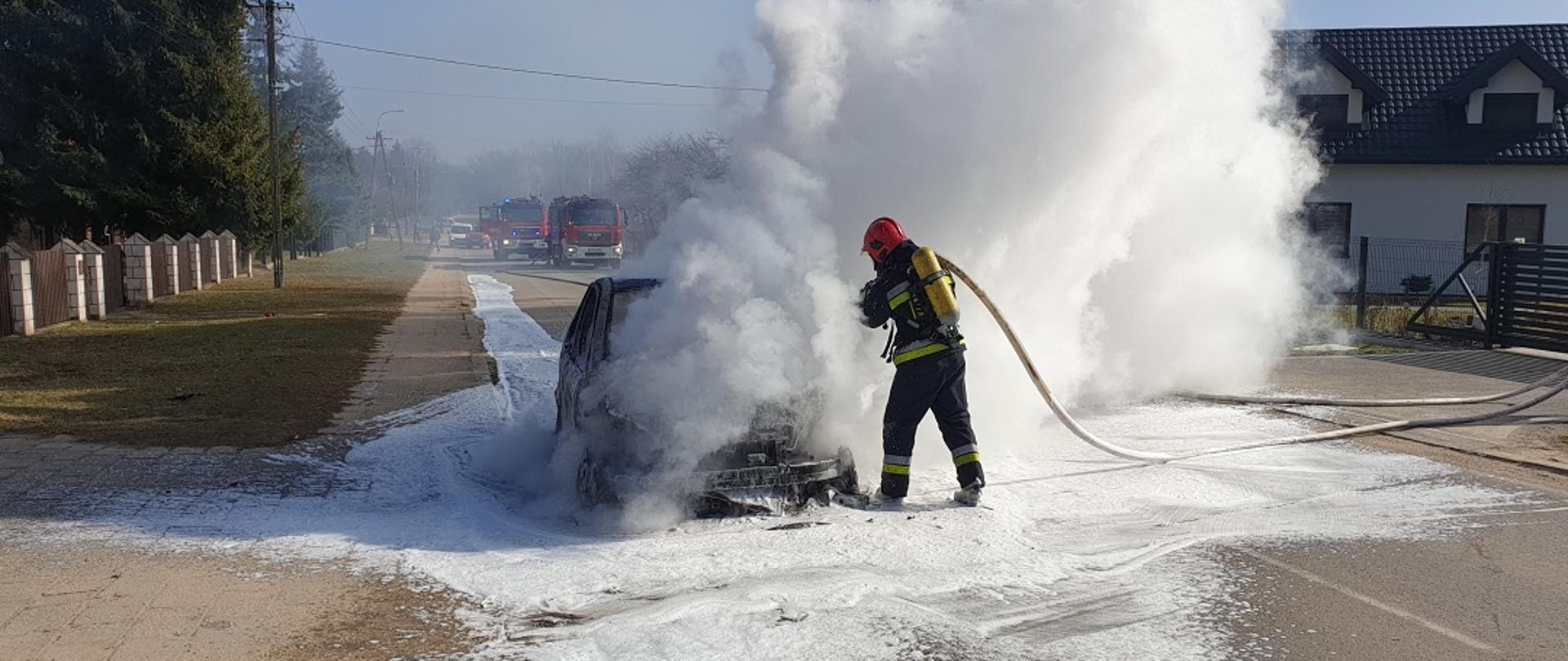 alternatywny: Na zdjęciu widzimy działania gaśnicze przy palącym się samochodzie. Strażacy zabezpieczeni w aparaty ochrony dróg oddechowych gaszą pojazd. W tle widać wozy strażackie. 