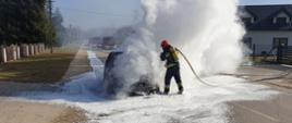 Na zdjęciu widzimy działania gaśnicze przy palącym się samochodzie. Strażacy zabezpieczeni w aparaty ochrony dróg oddechowych gaszą pojazd. W tle widać wozy strażackie. 