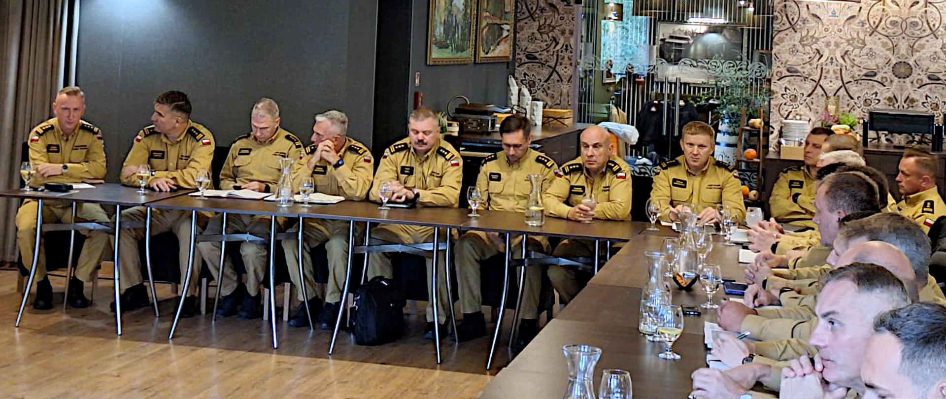 Na zdjęciu widać dużą salę podczas narady kadry kierowniczej państwowej straży pożarnej województwa śląskiego. Przy długim, złączonym w prostokąt stole siedzi kilkanaście osób -komendantów powiatowych i miejskich w identycznych, piaskowych mundurach strażackich. Siedzą w skupieniu, część notuje lub ma przed sobą dokumenty i szklanki z wodą. Na suficie wiszą duże, okrągłe lampy z jasnymi abażurami, a na ścianach widać dekoracyjną tapetę w geometryczne wzory. 