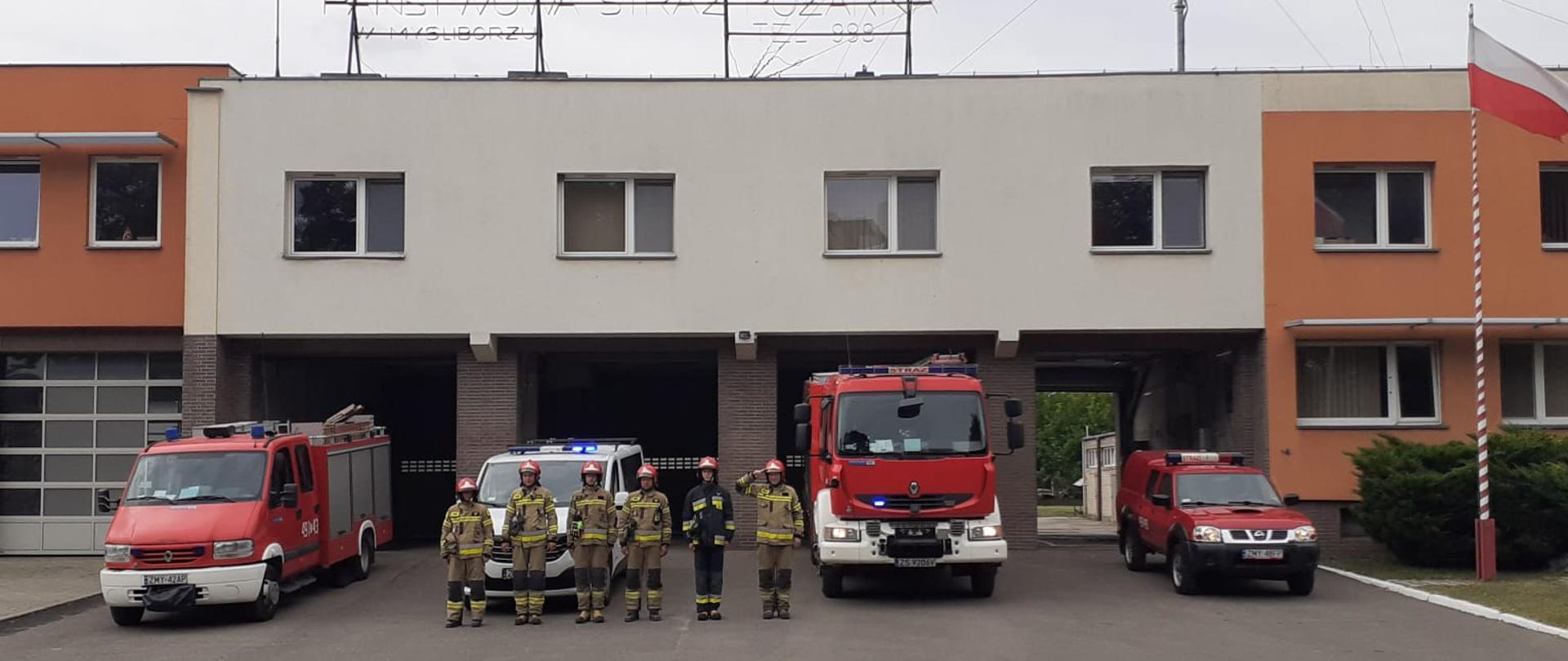 Strażacy stojący na baczność przed budynkiem komendy PSP. Za nimi samochody pożarnicze z włączoną sygnalizacją świetlną. Z prawej strony biało czerwona flaga powiewa na maszcie.