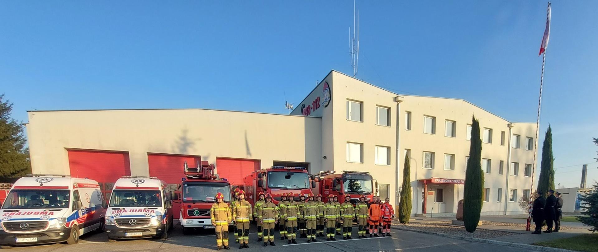 Plac przed budynkiem Komendy Powiatowej Państwowej Straży Pożarnej w Opocznie. Kilkunastu strażaków stojących w dwóch rzędach, wszyscy w ubraniach specjalnych z czerwonymi hełmami, obok stoi kilku ratowników medycznych w pomarańczowych ubraniach. Wszyscy w postawie zasadniczej na baczność. W tle trzy czerwone wozy bojowe strażackie oraz dwie karetki pogotowia ratunkowego.