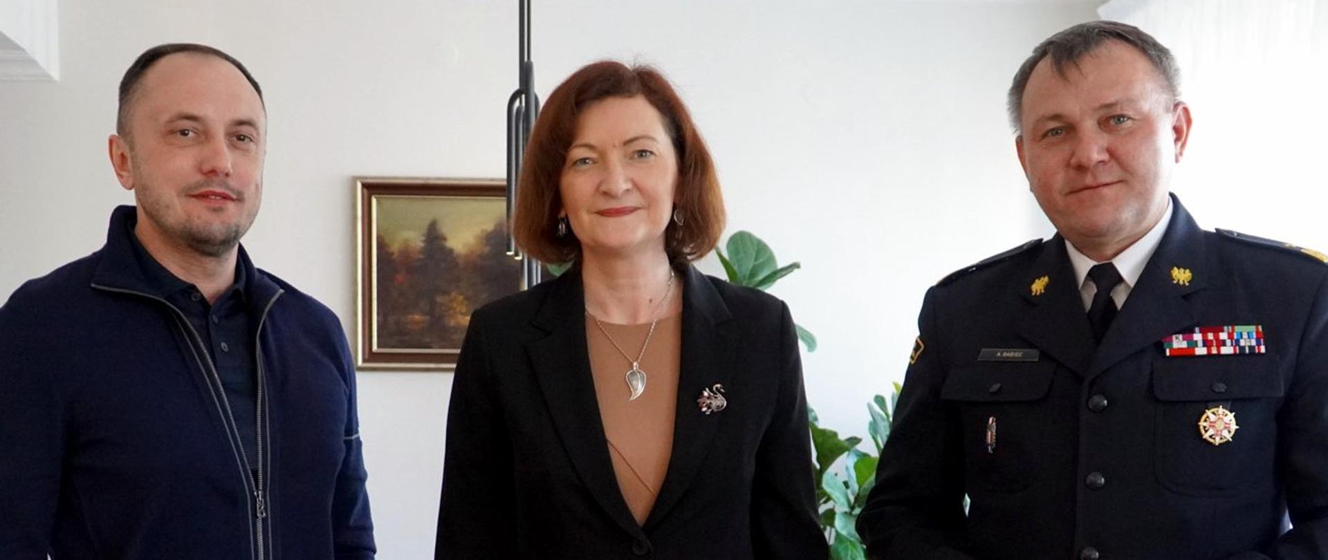 Zdjęcie zrobione w pomieszczeniu. Na fotografii w szeregu stoją trzy osoby, dwóch mężczyzn i kobieta. Jeden z mężczyzn to oficer Państwowej Straży Pożarnej w stopniu generała.