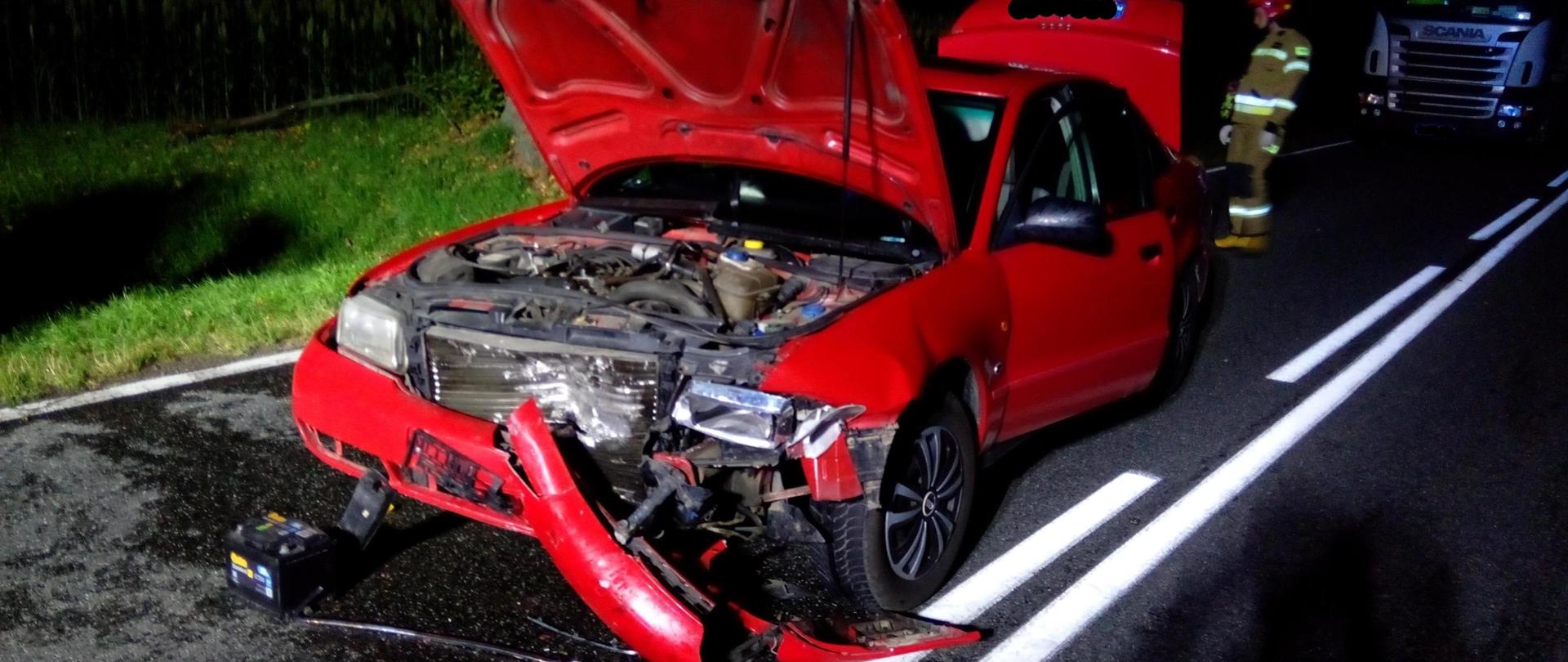 Czerwone auto z rozbitym przodem, w tle umundurowany strazak