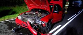 Czerwone auto z rozbitym przodem, w tle umundurowany strazak