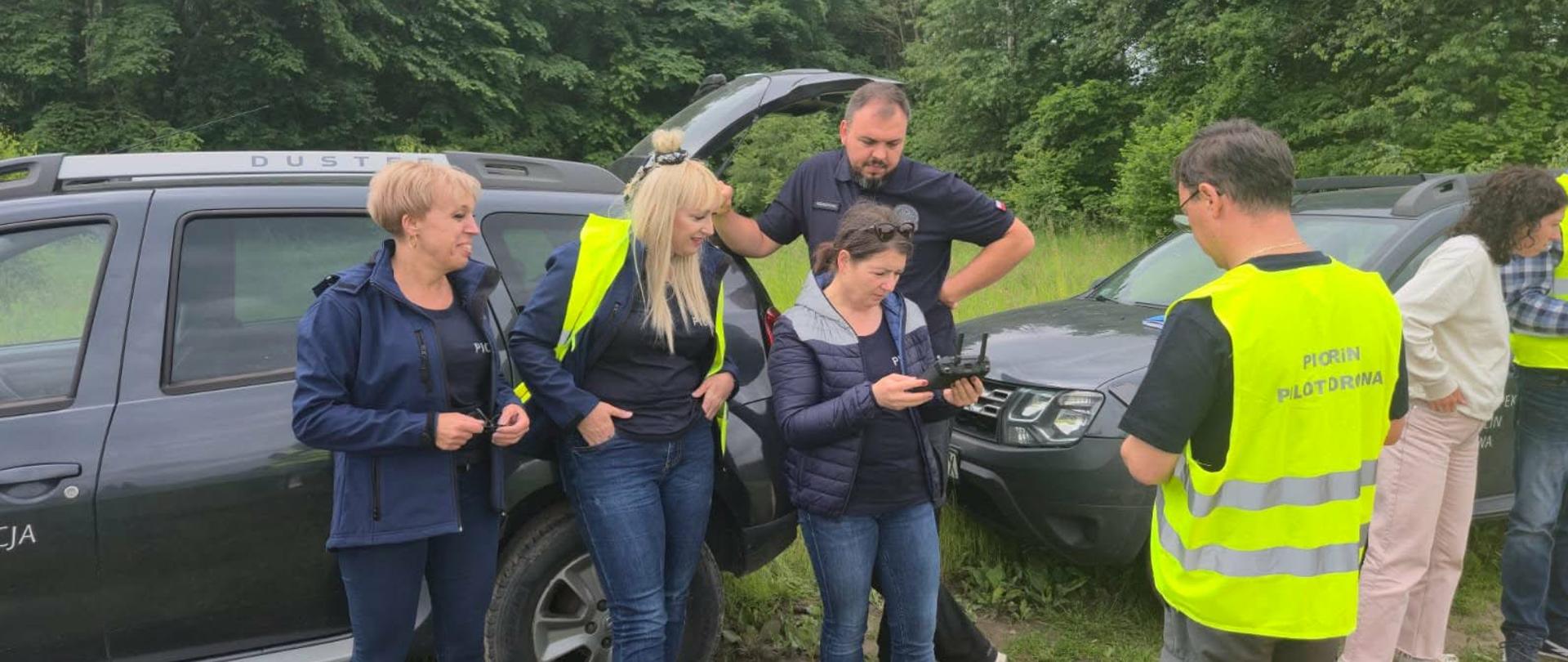 Grupa ludzi stoi przy samochodzie służbowym. Kobieta trzyma kontroler do drona