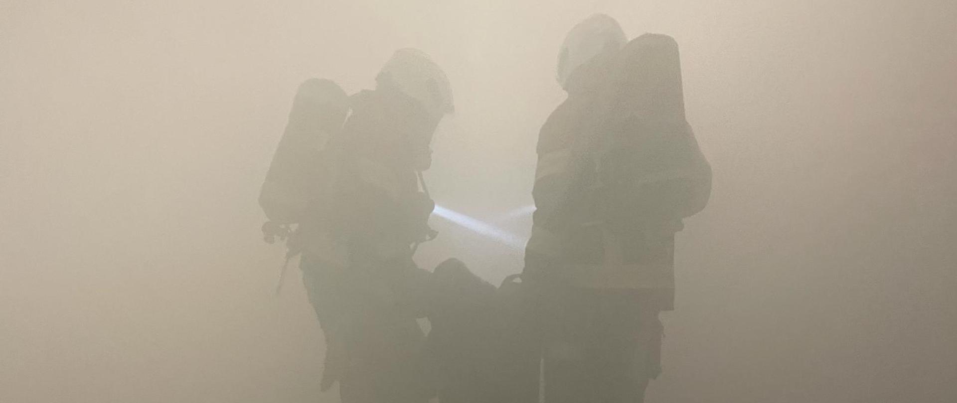 Zdjęcie przedstawia strażaków ochotników podczas ćwiczenia inspekcyjnego,ewakuujących poszkodowanego w gęstym zadymieniu