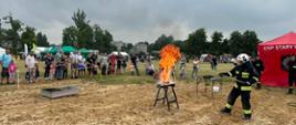 Na zdjęciu znajduje się strażak ubrany w "nomex", na stoliku stoi naczynie z którego bucha ogień, w tle widać wiele osób oraz drzewa i namiot OSP Stary Ujazd