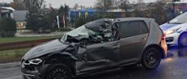 Zdjęcie przedstawia samochód osobowy koloru szary metalik ze zniszczonym lewym bokiem po wypadku. Za samochodem stoi radiowóz policyjny. 