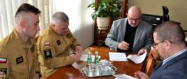 Przy stoliku na którym znajdują się dokumenty siedzą strażacy w piaskowych mundurach oraz mężczyźni w garniturach. W tle baner powiatu tarnowskiego i okna