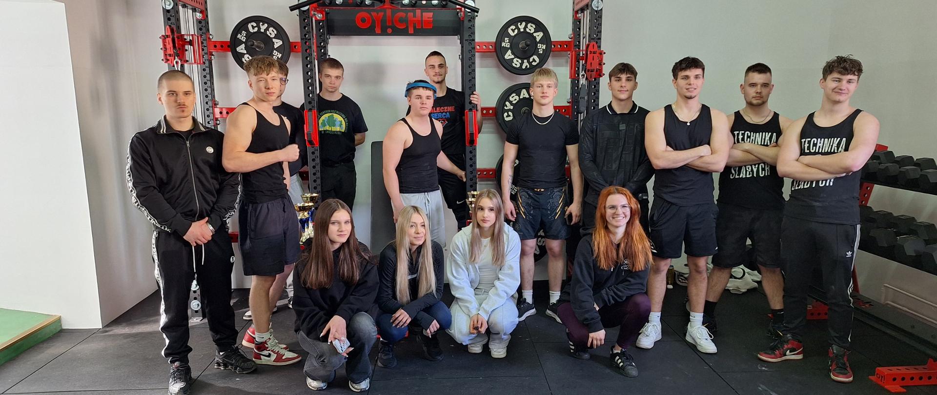 Grupa kilkunastu młodych osób, kobiet i mężczyzn w strojach sportowych, pozuje do wspólnego zdjęcia w sali gimnastycznej. Stoją przed profesjonalną klatką do ćwiczeń z napisem „OY!CHE”. Niektórzy zawodnicy mają założone pasy treningowe, w tle widoczne puchary i hantle.