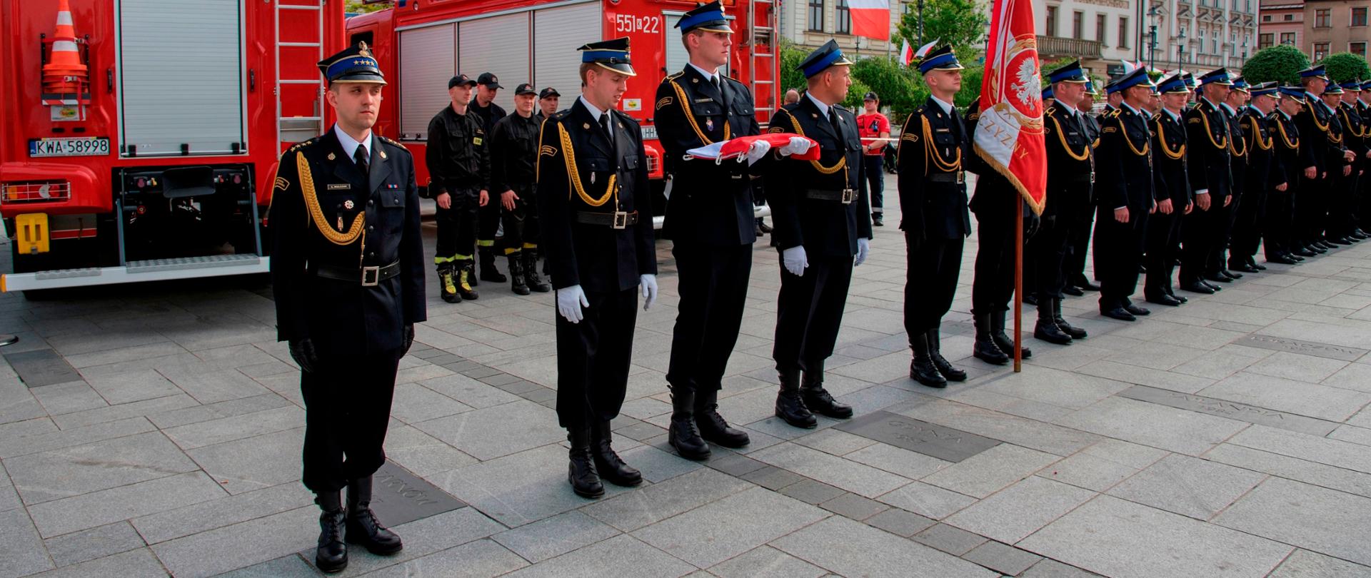 Widok z przodu. Poczet flagowy, poczet sztandarowy oraz pododdziały strażackie w szyku w trakcie uroczystości.