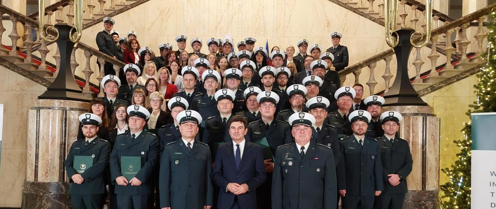Duża grupa osób w eleganckich, ciemnych mundurach służbowych stoi na reprezentacyjnych schodach na klatce schodowej w zabytkowym budynku. Uczestnicy, rozmieszczeni na schodach i podeście, noszą czapki służbowe, trzymają dyplomy. Na dole na czerwonym dywanie stoi kilku funkcjonariuszy oraz mężczyzna w garniturze. Po bokach schodów widoczne są ozdobne balustrady i kolumny a obok stoi ustrojona świąteczna choinka. Cała grupa pozuje do oficjalnego uroczystego zdjęcia.