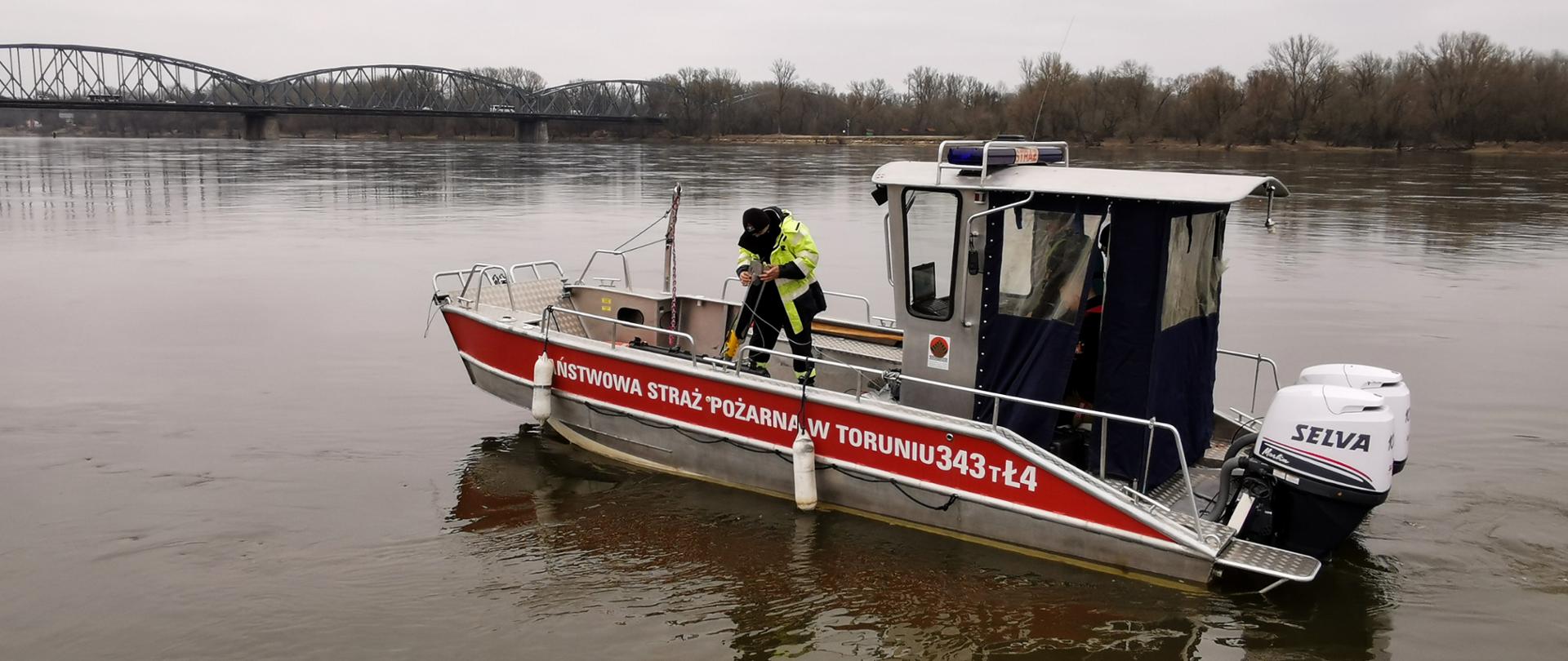 Strażak znajdujący się na łodzi przygotowuje sonar do uruchomienia. Łódź znajduje się na rzece Wiśle. W tle widać most. 