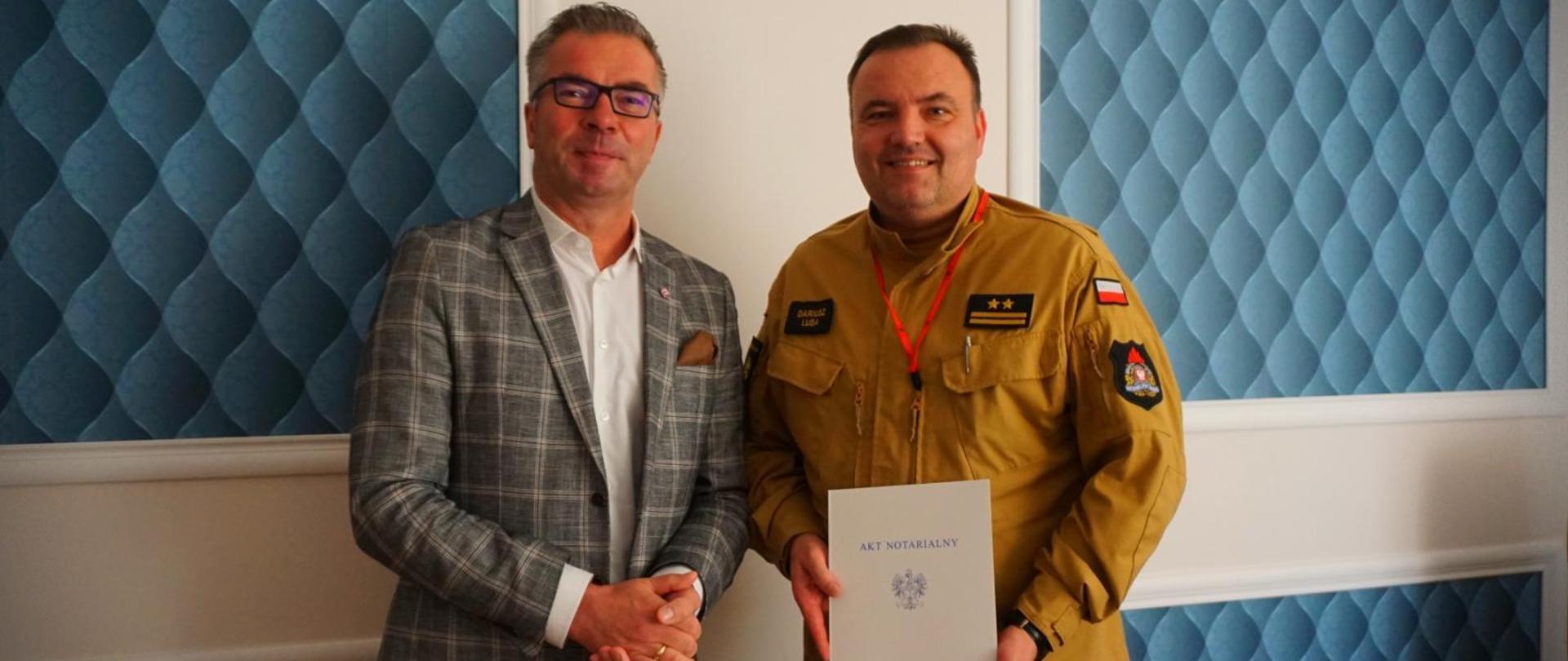 Prezydent Miasta Kutno oraz Komendant Powiatowy PSP w Kutnie na tle niebieskiej sztukaterii pozują do zdjęcia. Komendant Powiatowy PSP w Kutnie trzyma w dłoni Akt Notarialny oprawiony w białej teczce.