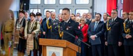 Na zdjęciu przy mównicy przemawia Komendant Powiatowy Państwowej Straży Pożarnej w Gorlicach podczas apelu z okazji Dnia Strażaka 2025. W tle zaproszeni na uroczystość goście. 