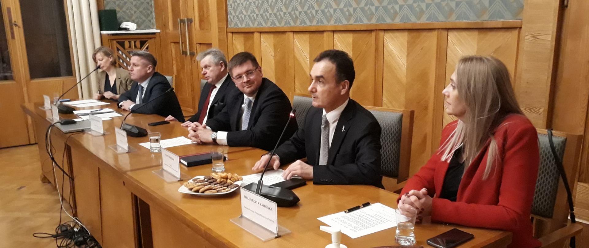 Grupa osób siedzi przy podłużnym stole, na pierwszym planie kobieta w czerwonym żakiecie, po środku siedzi wiceminister Rzymkowski. 