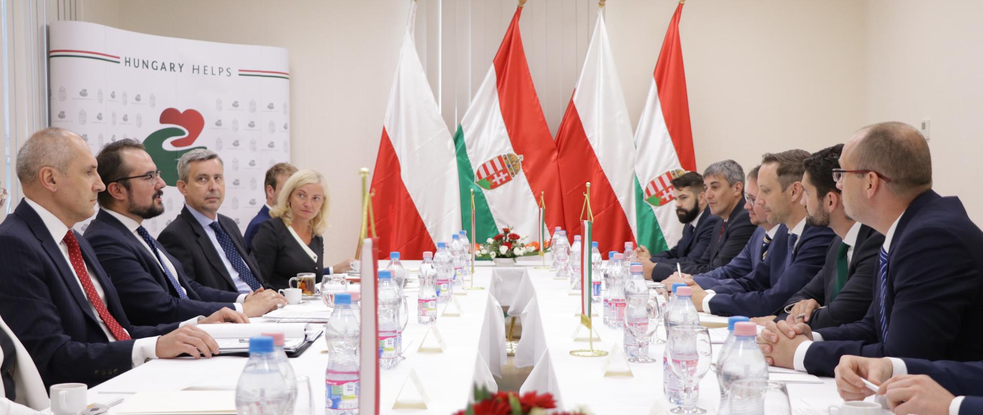 Group of people are sitting at the table. On the background there are Polish and Hungarian flags