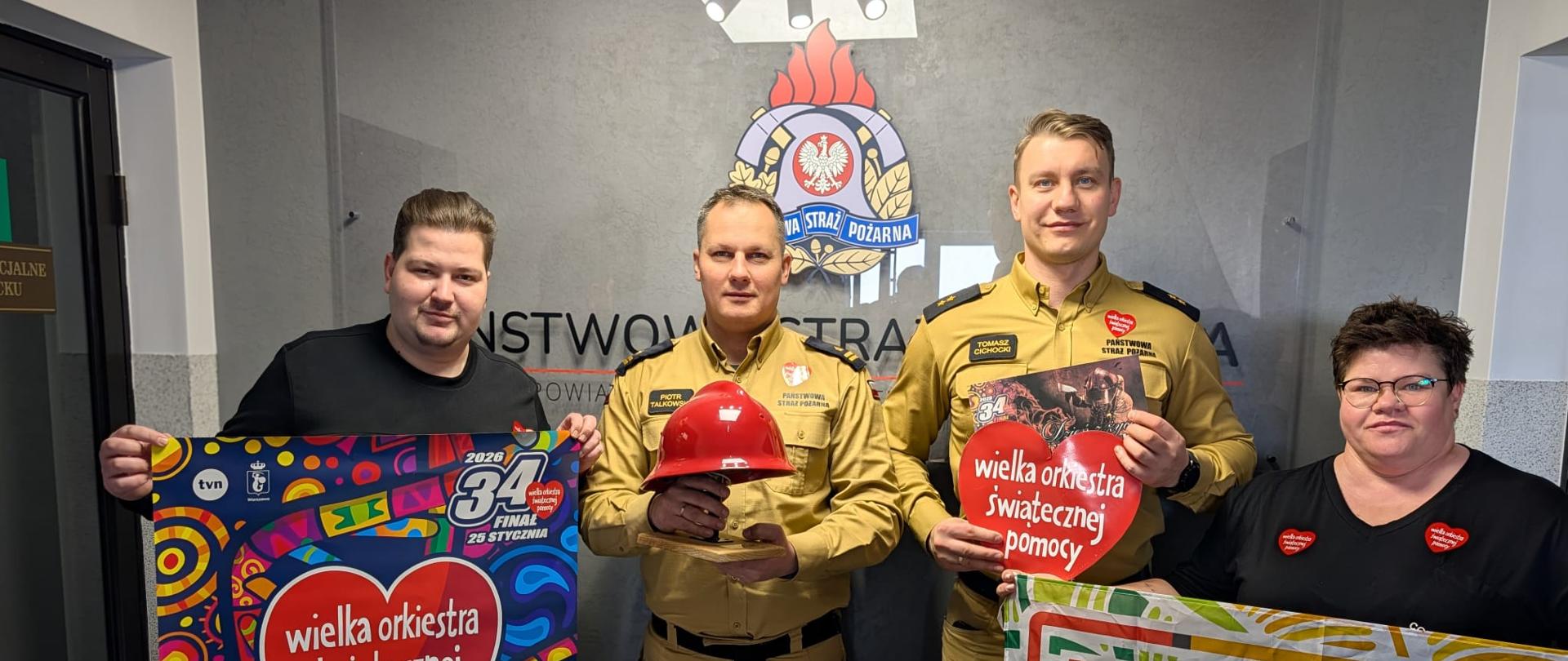 Zdjęcie przedstawia cztery osoby, 3 mężczyzn oraz kobietę. 2 mężczyzn to strażacy w umundurowaniu służbowym, kolejne dwie osoby to przedstawiciele sztabu WOŚP. Osoby trzymają w dłoniach przedmioty związane z Wielką Orkiestrą Świątecznej Pomocy.