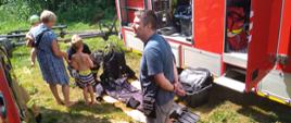 Pokaz sprzętu do nurkowania przy samochodach specjalnych na wyposażeniu Jednostki ratowniczo -Gaśniczej w Łomży. Strażak trzyma w ręku maskę pełną nurkową ,pokazuje małemu chłopcu który stoi przy swojej babci. Osoba dorosła przygląda się wyposażeniu samochodu gaśniczego. 