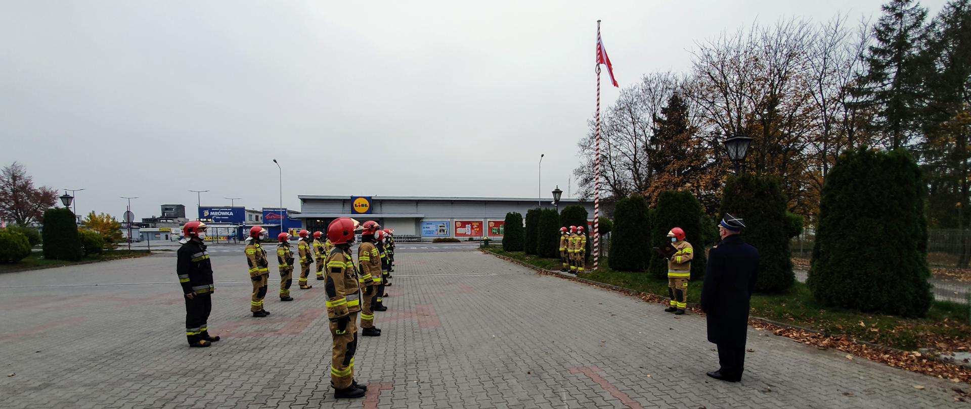 Strażacy stojący w dwuszeregu podczas uroczystej zbiórki na placu zewnetrznym komendy. W tle powiewająca flaga na maszcie. 