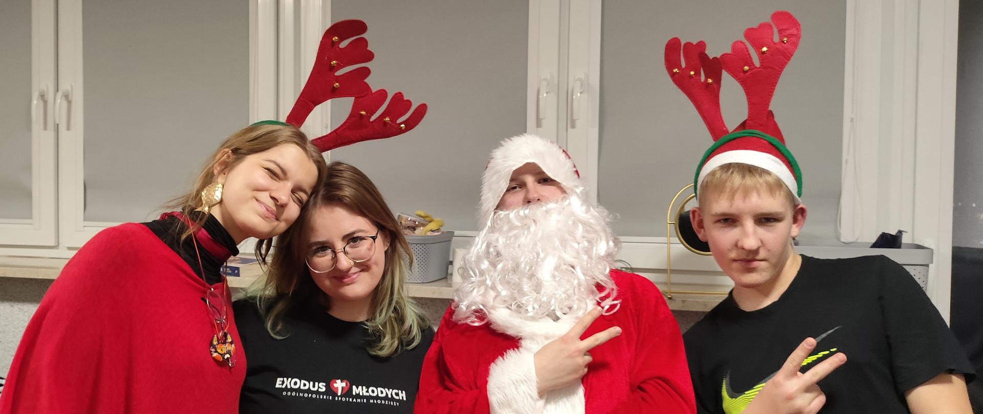 Czworo młodych ludzi pozuje do zdjęcia. Pośrodku jest chłopiec w stroju Mikołaja. Z prawej strony w czapce mikołajowej i czerwonych rogach renifera. Obok Mikołaja jest dziewczyna w czarnej bluzce, a lewej dziewczyna ubrana na czerwono i z czerwonymi rogami renifera na głowie. W tle widać szerokie okno osłonięte szarymi roletami.