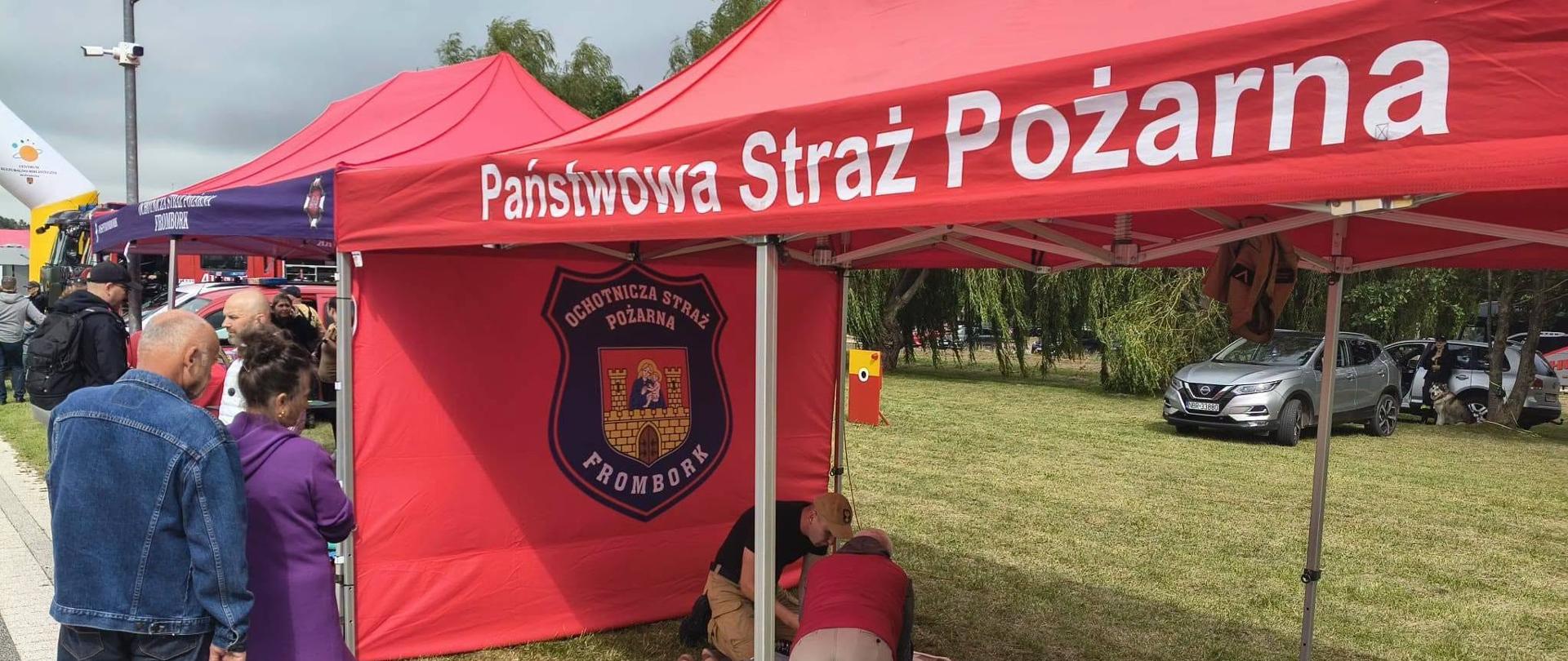 Na zdjęciu widać stoisko promocyjno-edukacyjne straży pożarnej podczas wydarzenia plenerowego. Pod czerwonym namiotem z napisem „Państwowa Straż Pożarna” prowadzone są pokazy z zakresu pierwszej pomocy. Na matach znajdują się fantomy do nauki resuscytacji krążeniowo-oddechowej (RKO), a uczestnicy ćwiczą pod okiem strażaka. Obok namiotu przechodzą odwiedzający, a w tle widoczne są kolejne stoiska oraz zaparkowane pojazdy.