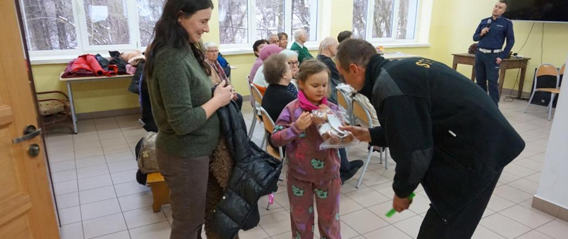 Strażak wręcza dziewczynce misia w tle uczestnicy spotkania