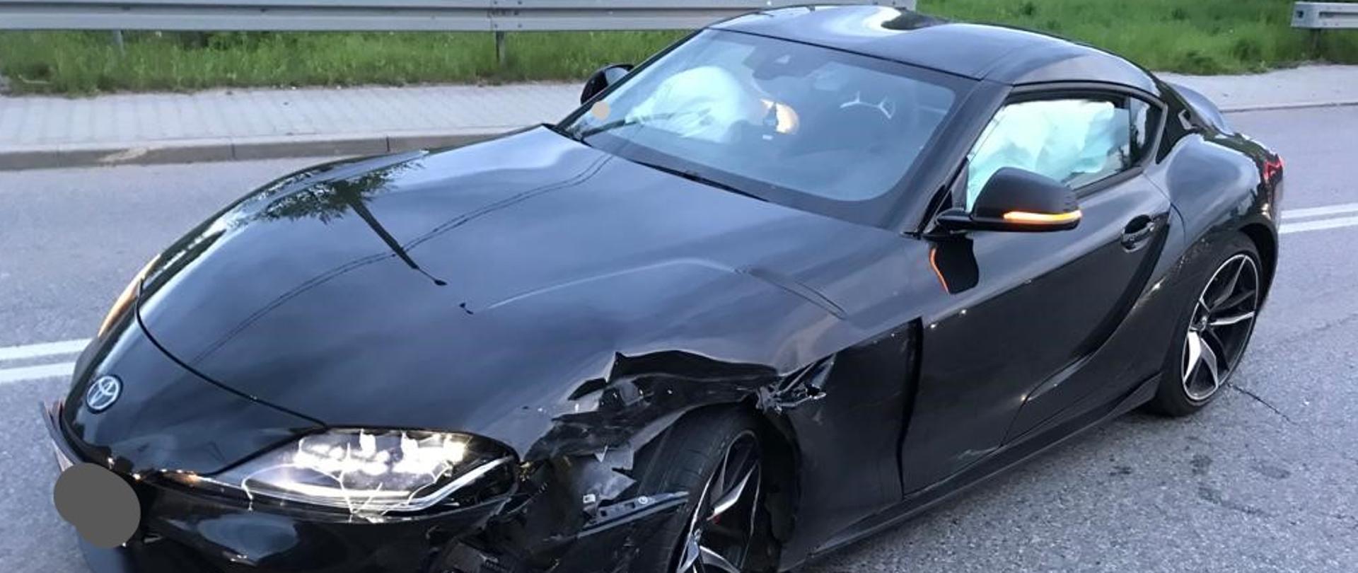 Na zdjęciu widzimy rozbity samochód osobowy w kolorze czarnym stojący na jezdni drogi wojewódzkiej. Za nim widoczne są zabudowania miejscowości. 