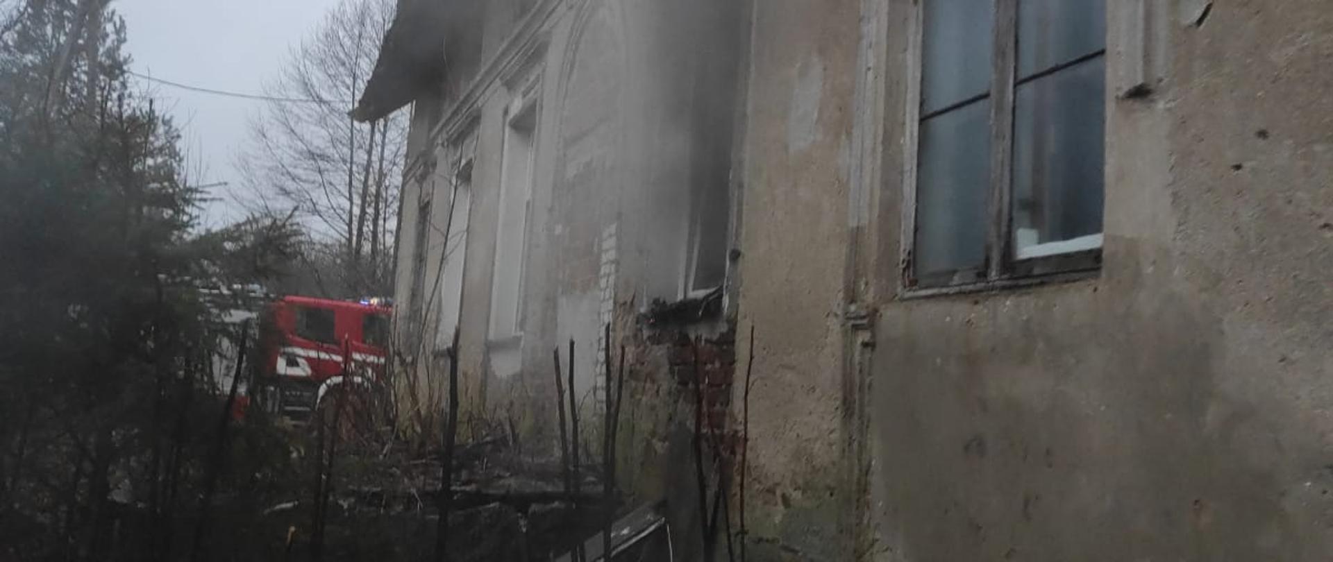 Z prawej strony szary budynek z którego wydobywa się dym. Dalej w oddali czerwony samochód strażacki