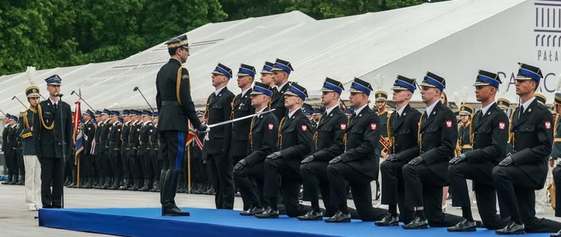 Zdjęcie przedstawia uroczysty moment promocji oficerskiej strażaków, odbywający się na świeżym powietrzu, na specjalnym podwyższeniu wyłożonym niebieskim dywanem. W prawym przednim rzędzie na baczność, klęcząc na jednym kolanie, stoi grupa kilkunastu strażaków w czarnych mundurach wyjściowych, czapkach rogatywkach i czarnych rękawiczkach. Przed nimi, po lewej stronie, stoi tyłem generał , który dokonuje tradycyjnego pasowania szablą, dotykając nią ramienia jednego z klęczących funkcjonariuszy. W tle, po lewej stronie placu, widać długi szereg kolejnych umundurowanych strażaków stojących w rzędzie.