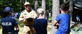 Na zdjęciu widzimy grupę pięciu osób stojących na zewnątrz w lesie, podczas kontroli obozu harcerskiego wykonywaną przez strażaków i policję. Na pierwszym planie po lewej stronie stoi kobieta w mundurze policyjnym z napisem „POLICJA” na plecach. Ma ciemnogranatowy mundur, pas z kaburą i kajdanki oraz czapkę z daszkiem. Na środku stoi mężczyzna, strażak w stopniu starszego kapitana w jasnobrązowym mundurze. Ma na głowie czapkę z daszkiem i trzyma w rękach czarna teczkę z dokumentami. Przed nim stoją tyłem dwie osoby w cywilnych ubraniach – kobieta z jasnymi włosami spiętymi w kok, w ciemnej bluzce i jeansowych spodenkach oraz mężczyzna w niebieskiej koszulce i czarnych spodenkach. W tle widać drzewa, rozwieszone kolorowe ubrania i namiot w których będą spać harcerze podczas obozu w lesie.