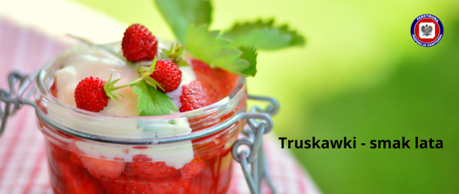 Na stoliku deser truskawkowy w słoiku przybrany śmietaną poziomkami i listkami mięty. W prawym górnym rogu logo Państwowej Inspekcji Sanitarnej. Na zielonym tle ciemny napis Truskawki - smak lata.