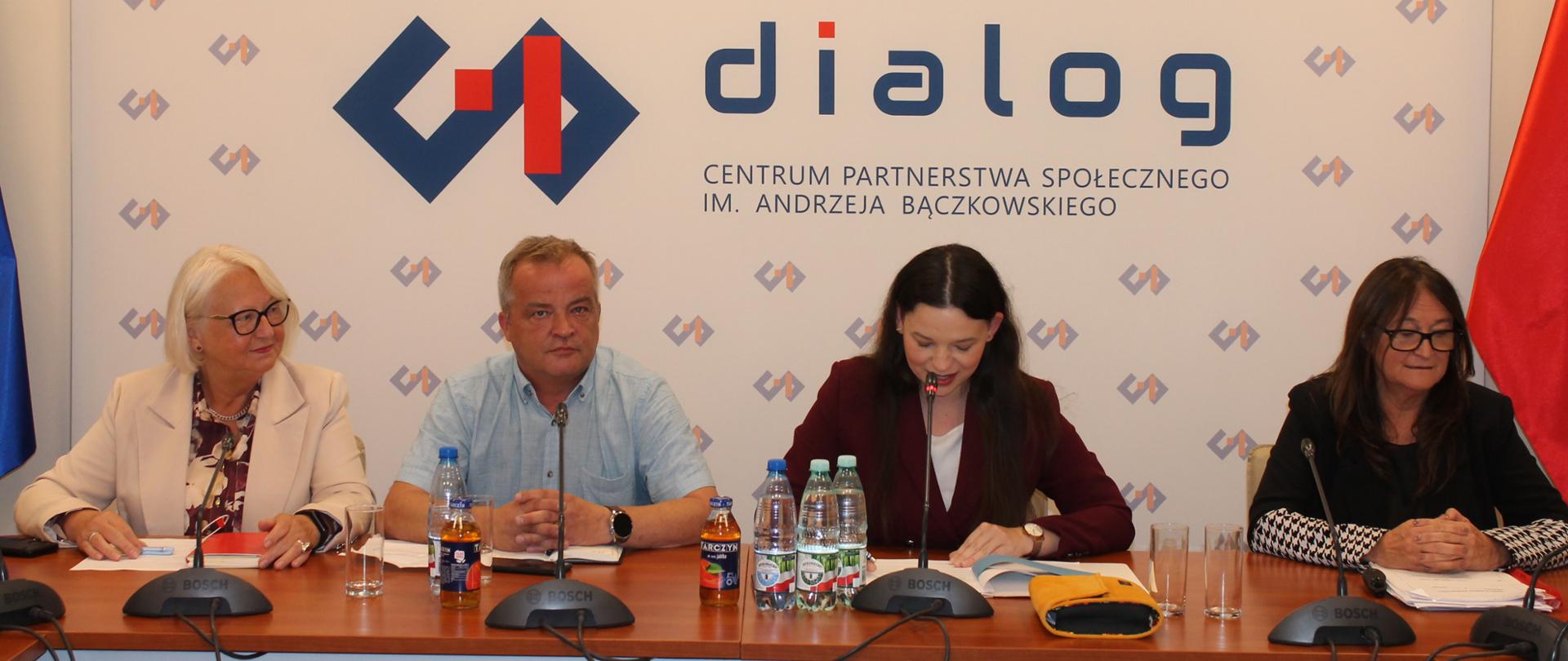 Na zdjęciu widnieje stół prezydialny za którym siedzi wiceminister Rodziny, Pracy i Polityki społecznej – Katarzyna Nowakowska, która prowadzi spotkanie. W tle znajduje się baner z napisem Centrum Partnerstwa Społecznego Dialog. Po lewej stronie Ministry siedzi Beata Karlińska Dyrektor Departamentu pomocy Społecznej, a po prawej stronie zasiadają współprzewodnicząca Zespołu ze strony pracodawców – Teresa Hernik oraz Zbigniew Spendel – współprzewodniczący strony związkowej. 