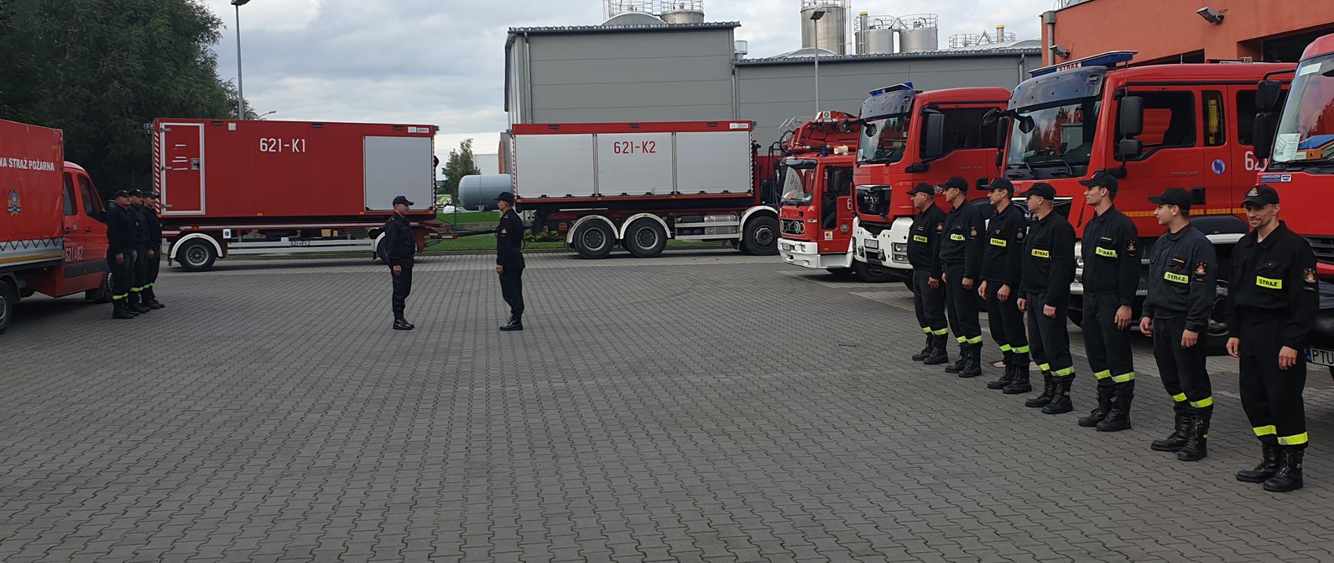 Dowódca JRG składa meldunek Zastępcy Komendanta Powiatowego. Strażacy stoją w szeregu przed pojazdami pożarniczymi.