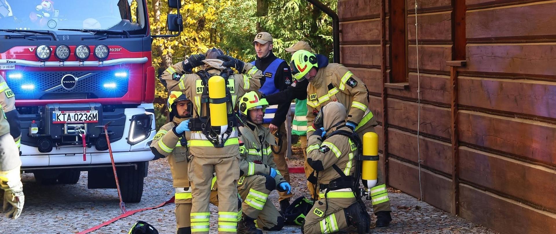 Grupa strażaków znajduje się obok drewnianego budynku. Część z nich ma ubrane aparaty ochrony dróg oddechowych. Większość z nich ubranych jest w piaskowe ubrania specjalne dodatkowo obok znajduje się wóz strażacki z uruchomionymi niebieskimi światłami 