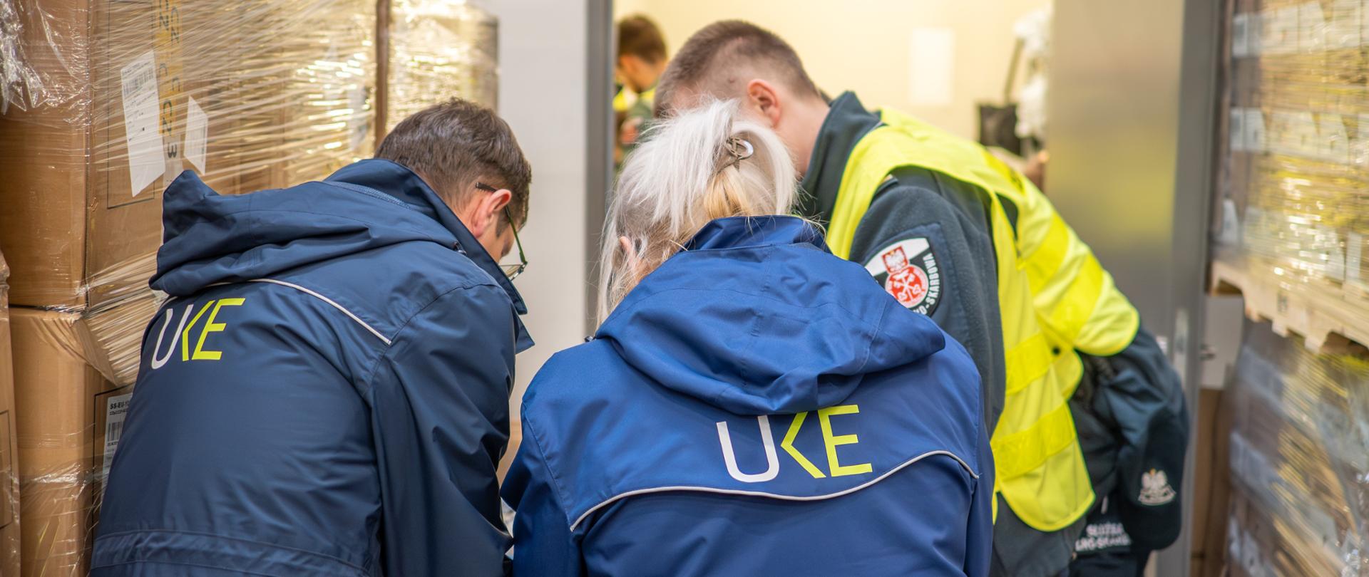 Trzy osoby w odzieży służbowej pochylają się nad stołem w magazynie, gdzie w tle znajdują się palety owinięte folią. Na plecach dwóch osób widoczny jest napis „UKE”, a obok stoi osoba w odblaskowej kamizelce.
