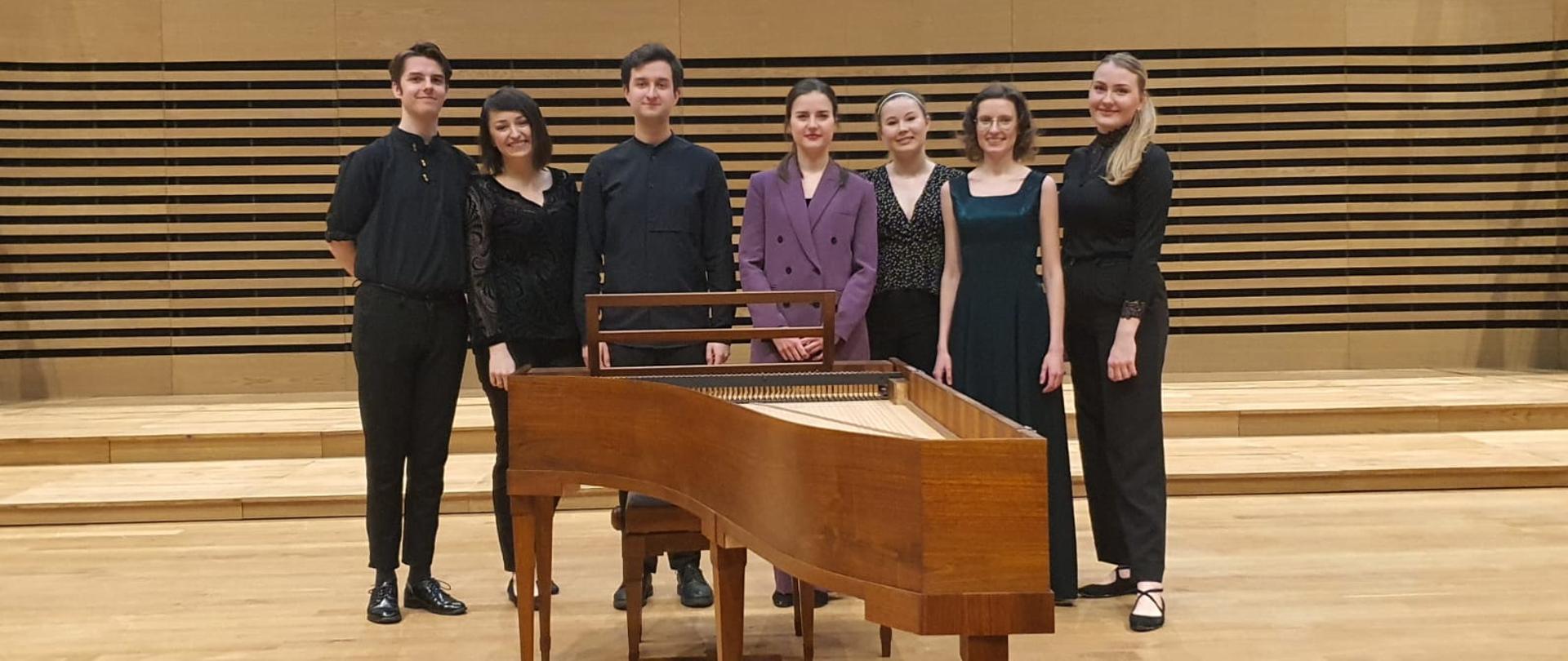 Dwóch mężczyzn i pięć kobiet stoi w rzędzie za fortepianem historycznym.