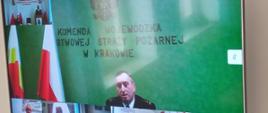 Na zdjęciu widoczny ekran telewizora na środku ekranu przemawiający małopolski komendant wojewódzki Państwowej Straży Pożarnej st. bryg. Piotr Filipek u dołu ekranu i po lewej stronie uczestniczący rocznej odprawy. 