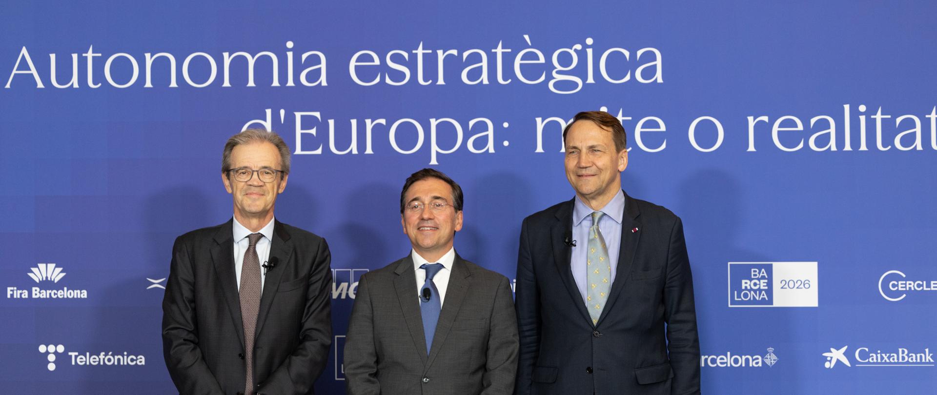 The photo shows three men in suits. They are looking toward the camera. Behind them is a blue wall with various white inscriptions. 