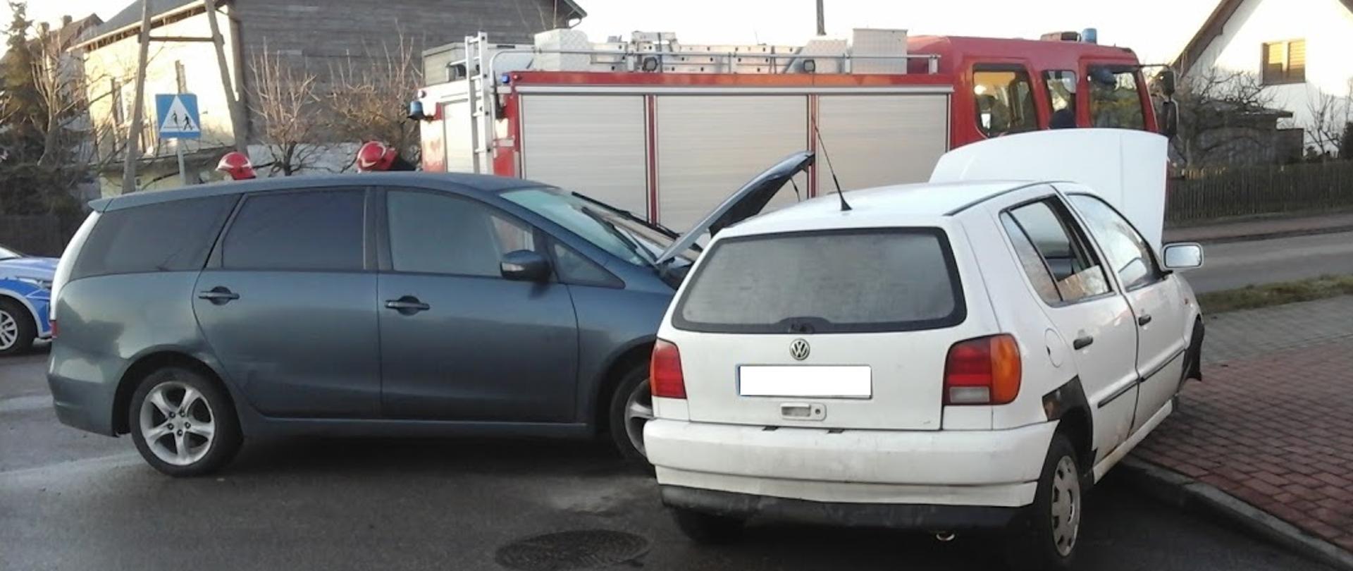Na zdjęciu widać rozbity dwa samochody .biały samochód osobowy znajdujący się na chodniku. W oddali wóz ratownictwa technicznego 
