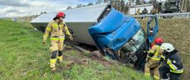 Zdjęcie przedstawia na pierwszym planie uszkodzone auto ciężarowe na poboczu drogi w rowie, po lewej i prawej stronie stojący funkcjonariusze PSP w hełmach i ubraniu tupu Nomex w trakcie działań. W tle pobocze drogi S10 i rów porośnięte trawą. W gór4nym prawym rogu zastęp i strazacy z WSP Wałcz na tle drogi S10 obwodnicy Wałcza. 