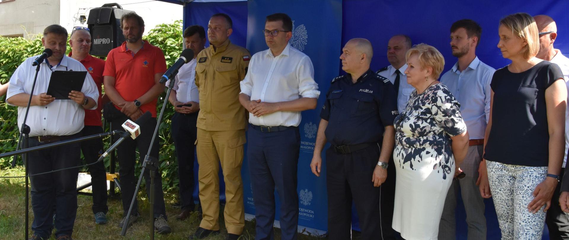 Przedstawiciele służb stoją pod niebieskim namiotem. Trwa briefing prasowy.