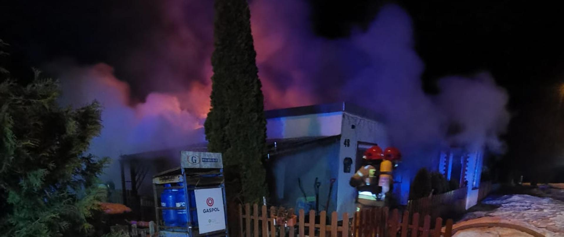 kadr przedstawie domek z płotkiem do kolan, za płotem soją strażacy w ubraniach specjalnych. Dom przesłania skrzynka z butlami z gazem propan-butan i wysoki krzew. Nad domem unosi się gęsty dym.