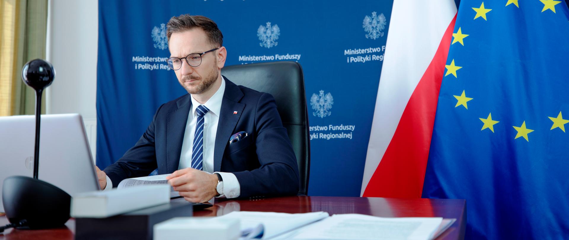 Wiceminister Waldemar Buda siedzi przed laptopem. W ręku trzyma dokumenty. Za nim ścianka promocyjna MFiPR oraz dwie flagi UE i PL.
