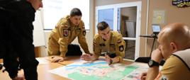 Kilku strażaków pochylają się nad stołem, czytają mapy, dyskutują gestykulując. Za nimi wyświetlane są obrazy na ekranie, z prawym górnym rogu fragment logo.