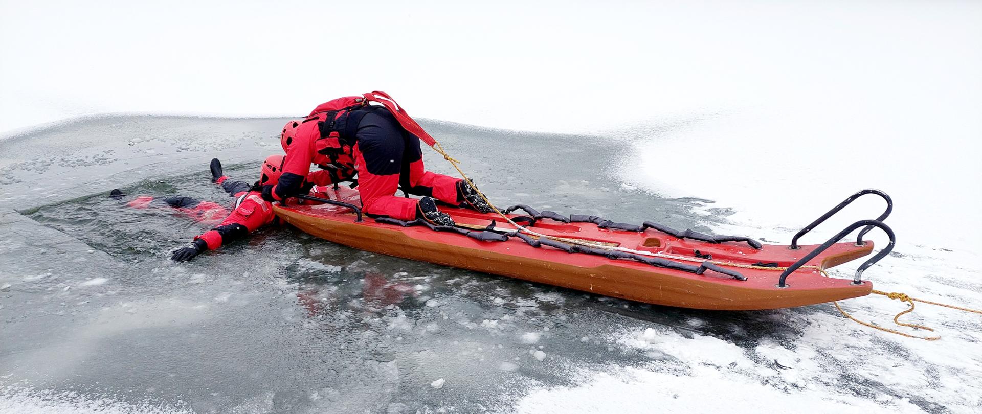 Zdjęcie przedstawia strażaka-ratownika znajdującego się na saniach lodowych, który
wyjmuje innego strażaka z przerębla.