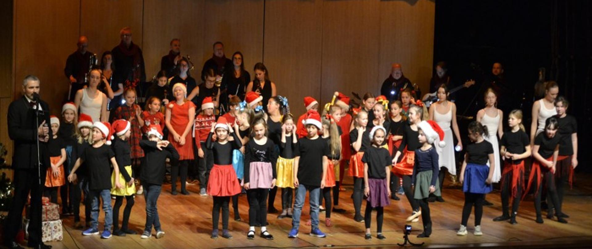 Zdjęcie kolorowe. Na scenie uczniowie w kolorowych strojach, orkiestra jazzowa i konferansjer.