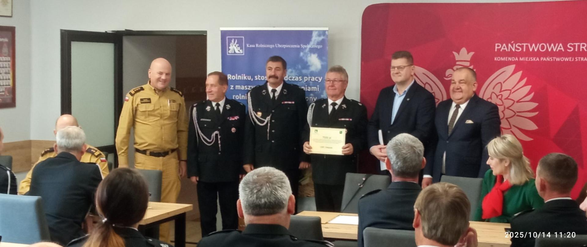 Na sali znajduje się grupa osób siedzących na krzesłach. Przed nimi stoją przedstawiciele OSP oraz KRUS, a za nimi znajduje się ścianka Państwowej Straży Pożarnej oraz roll-upy Kasy.