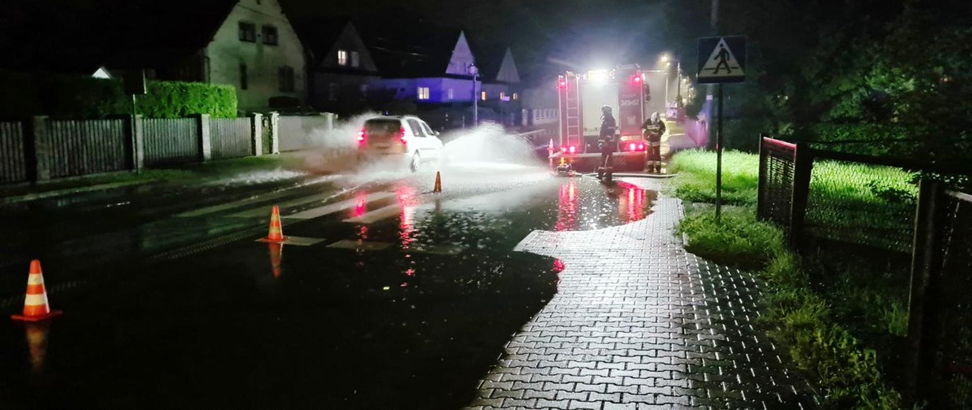 Rozlewisko na drodze oraz pachołki. Na poboczu stoi samochód pożarniczy a drogą jadzie samochód osobowy.