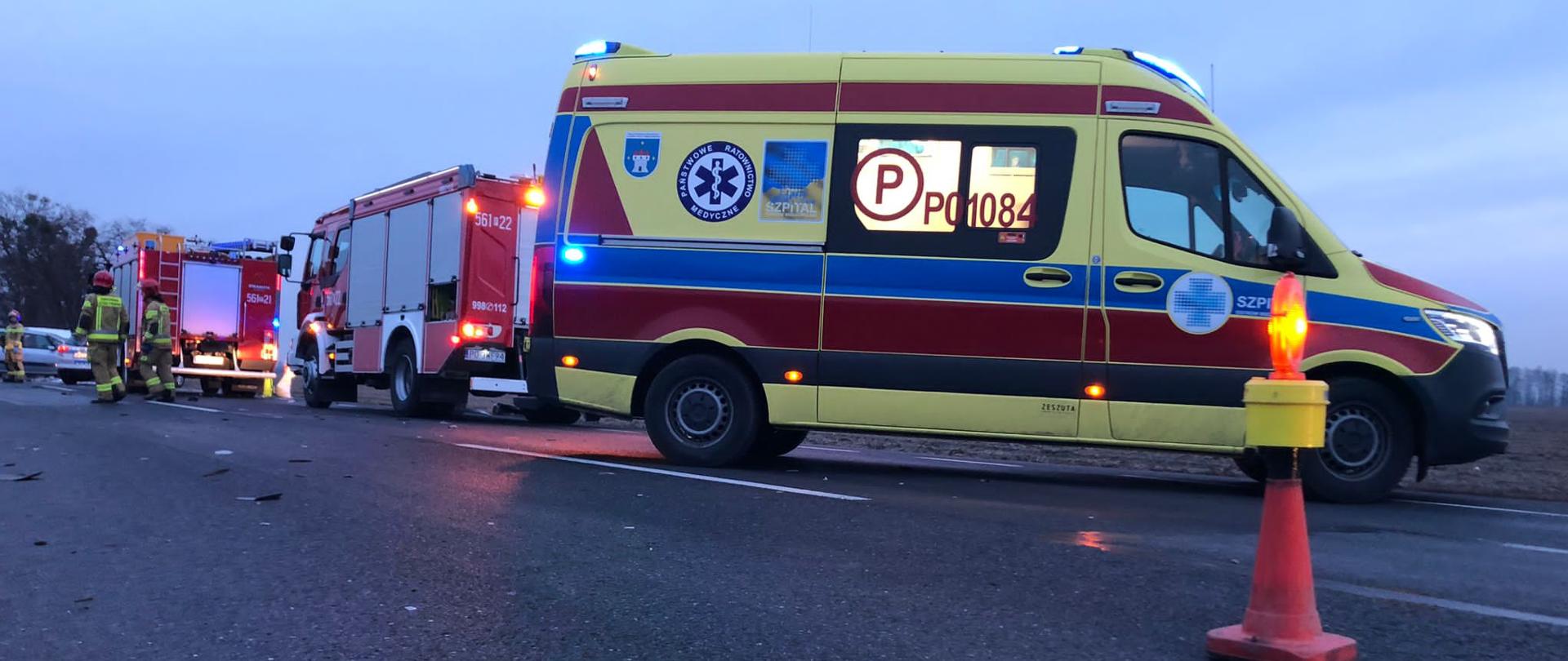 Jezdnia, ne pierwszym planie po prawej stronie pachołek ostrzegawczy z lampą błyskową, za nim stoi ambulans, dalej dwa wozy strażackie przy których widać strażaków